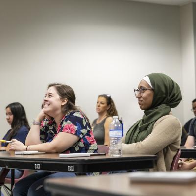 A classroom setting with students attentively listening to a lecture. The foreground features a student wearing a green hijab and glasses, smiling as she engages in the lesson. Other students are visible in the background, contributing to the atmosphere of focused learning.